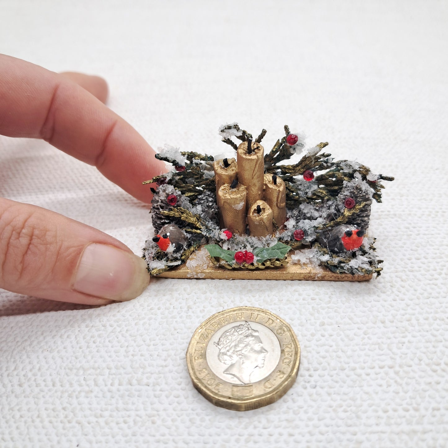 a dolls house miniature Christmas table centrepiece with gold candles, robins and artificial snow.. The miniature is next to a pound coin and hand for scale.