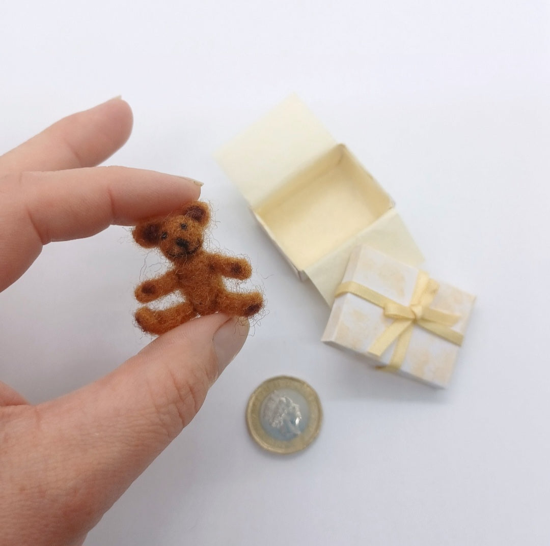 A dolls house teddy bear with gift box. The bear is light brown and felted from merino wool. A hand is holding the teddy bear for scale purposes.