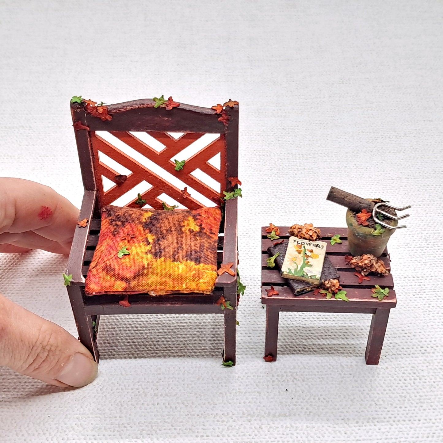 A dolls house miniature garden table and chair set in brown and burnt orange decorated with Autumnal leaves and garden accessories. A hand is shown next to the chair for scale. 