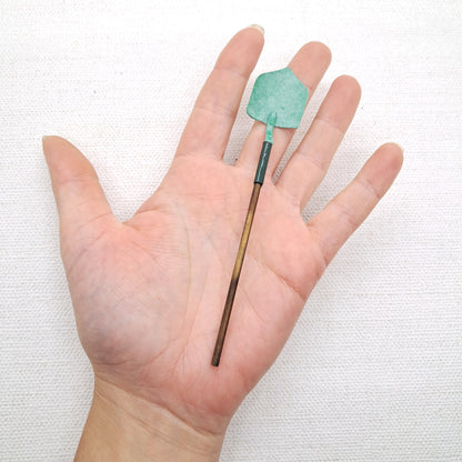 A hand is holding a long handled garden spade for dolls houses in the miniature 12th scale. The spade in sage green and brown.