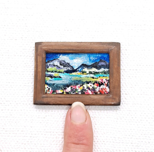 A miniature framed painting. A landscape painting that is acrylic on canvas housed in a brown wooden frame. A finger is shown next to the painting for scale.