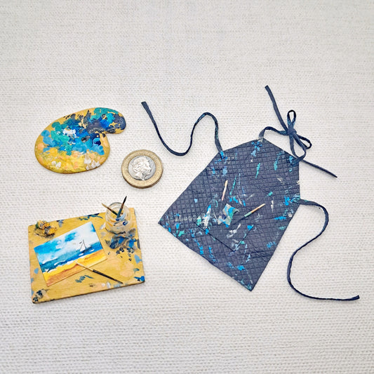 A dolls house miniature artist set containing a navy blue leather apron miniature paint palette and artists board with painting and brush jar.