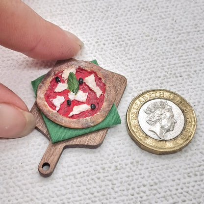 Dolls house miniature pizza on a chopping board. The pizza is margherita with black olives and basil leaf. It is hand painted polymer clay and measures 1 inch. It is next to a pound coin and a hand for scale.