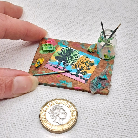 A tiny artists set on a wooden board. Miniature 1 inch landscape painting with a glass brush jar, paint brushes, a paint palette and cleaning rags. For 12th scale miniature artists studios