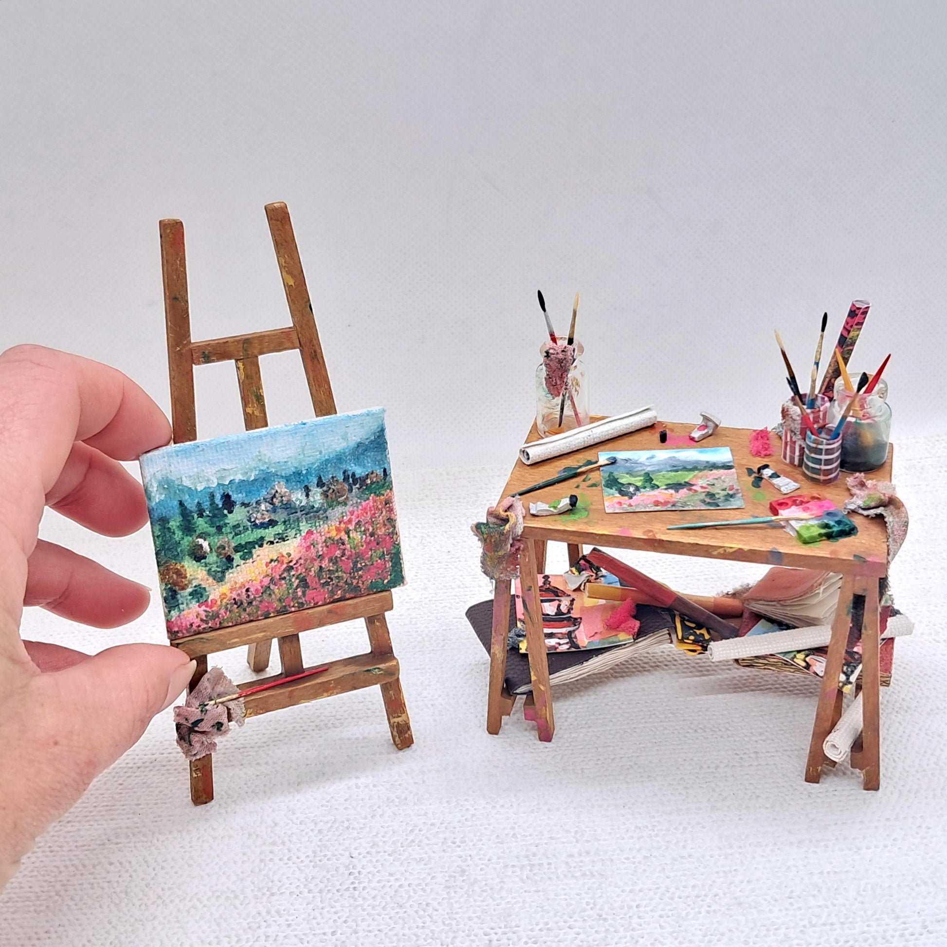 A 12th scale dolls house miniature wooden easel, canvas painting and accessorised wooden artists desk. 