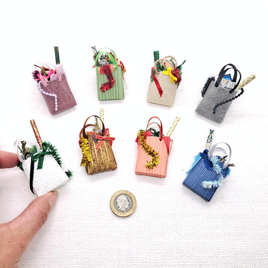 a hand is shown next to a group of 8 miniature Christmas shopping bags. The bags are for dolls house or miniature application and each contain Christmas colours, tinsel, gift wrap, and a tiny present.