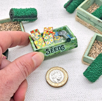 dolls house miniature green garden tray with seeds painted on the front. It contains seed packets and a garden trowel. A hand is holding it for scale purposes.