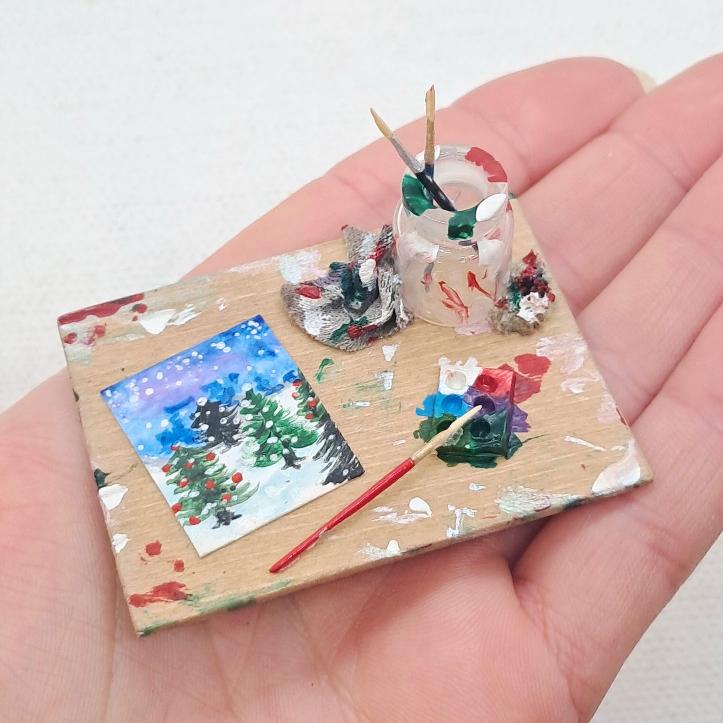 A dolls house miniature Christmas tree painting on a wooden board with a glass paint brush jar, tiny paintbrushes, a paint palette and cleaning cloths. A hand is holding the board for scale purposes. 